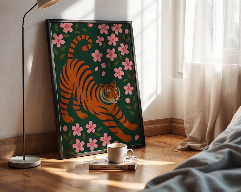 May include: Framed art print featuring an orange tiger with black stripes, stretching amidst pink cherry blossoms on a dark green background. The artwork is in a black frame and leans against a wall. A coffee cup and book are in front of the artwork.