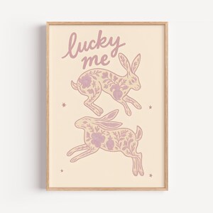 May include: A framed art print featuring two running rabbits with floral patterns in a soft pink hue. The words "lucky me" are written above the rabbits in a cursive font. The background is a light beige color.