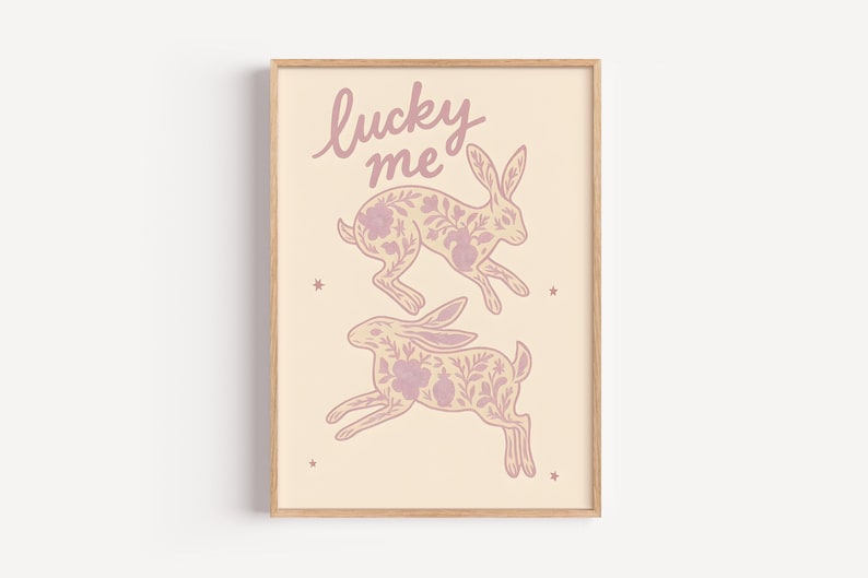 May include: A framed art print featuring two running rabbits with floral patterns in a soft pink hue. The words "lucky me" are written above the rabbits in a cursive font. The background is a light beige color.