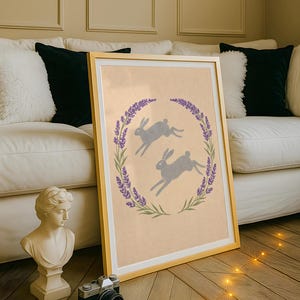 May include: A framed print with a gold frame, depicting two grey hares leaping within a lavender wreath. The artwork has a light beige background. The print is leaning against a white sofa with black and white cushions.