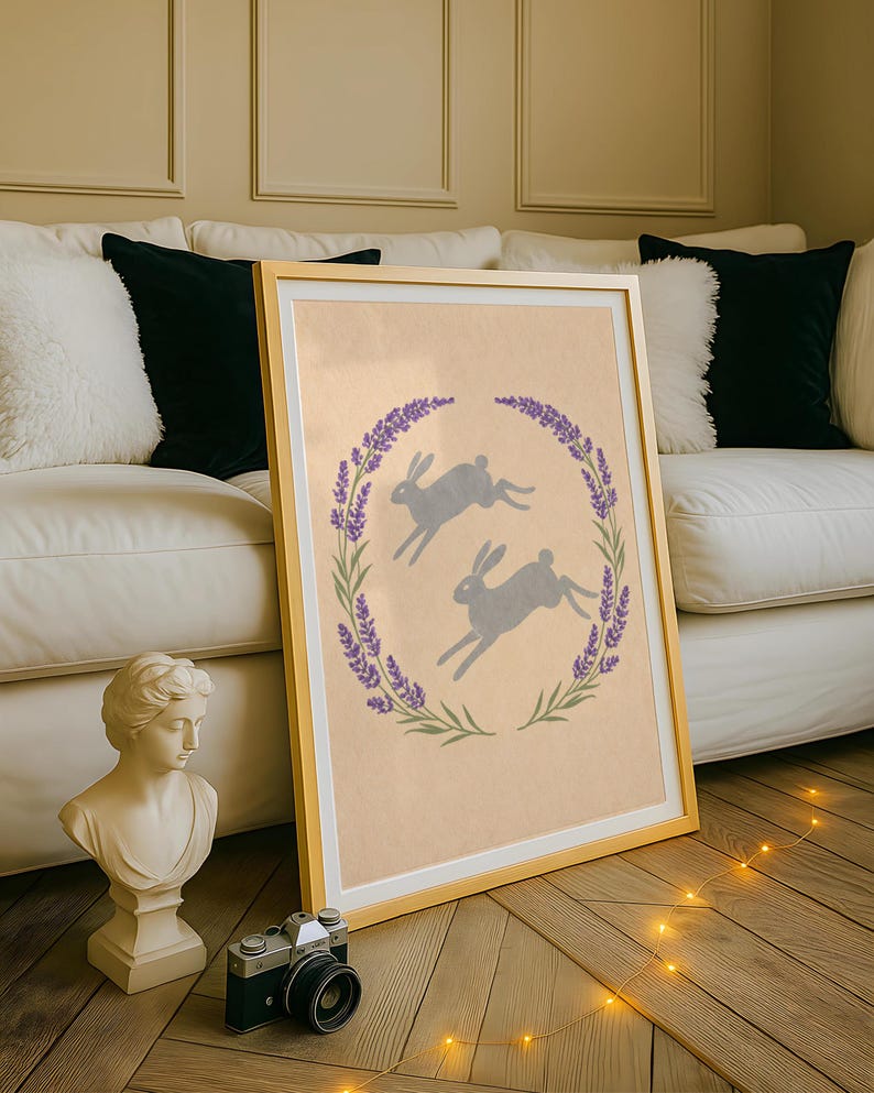 May include: A framed print with a gold frame, depicting two grey hares leaping within a lavender wreath. The artwork has a light beige background. The print is leaning against a white sofa with black and white cushions.