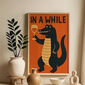 May include: A framed art print with a cartoon alligator holding a wine glass, set against an orange background. The text "IN A WHILE" is displayed at the top. The print is in a wooden frame.