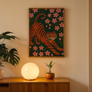 May include: A framed art print featuring an orange tiger with black stripes, set against a dark green background with pink floral accents. The artwork is displayed on a light-colored wall above a wooden cabinet. A round, glowing lamp and potted plants are in the foreground.