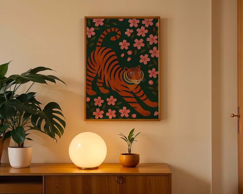 May include: A framed art print featuring an orange tiger with black stripes, set against a dark green background with pink floral accents. The artwork is displayed on a light-colored wall above a wooden cabinet. A round, glowing lamp and potted plants are in the foreground.