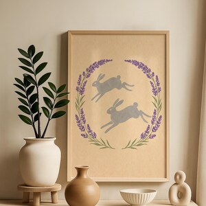 May include: A framed print with two grey hares leaping inside a lavender and green wreath on a tan backdrop. The print is displayed on a light wooden surface, with a white vase and greenery, a brown vase, a white bowl, and a decorative sculpture.