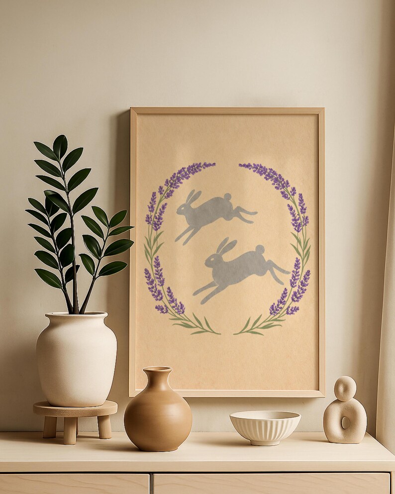 May include: A framed print with two grey hares leaping inside a lavender and green wreath on a tan backdrop. The print is displayed on a light wooden surface, with a white vase and greenery, a brown vase, a white bowl, and a decorative sculpture.
