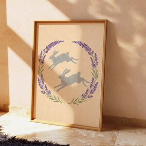 May include: A framed print showing two grey hares leaping inside a lavender and green floral wreath. The artwork is set against a beige background, with a wooden frame. The theme is nature and springtime.