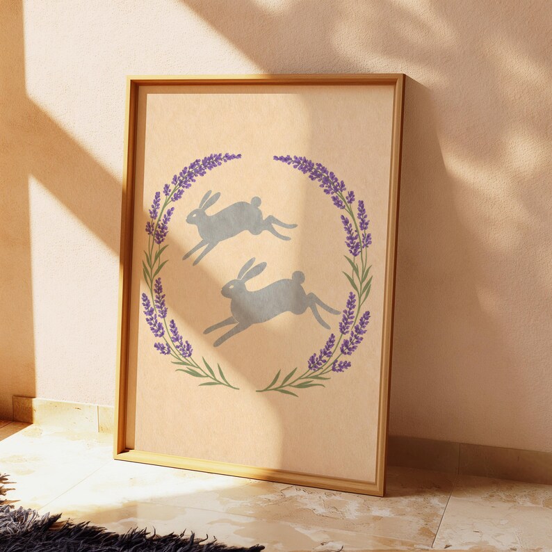 May include: A framed print showing two grey hares leaping inside a lavender and green floral wreath. The artwork is set against a beige background, with a wooden frame. The theme is nature and springtime.