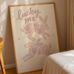 May include: A framed art print featuring two running rabbits with floral patterns in a soft purple hue. The words "lucky me" are written above the rabbits in a cursive font. The background is a light beige color.