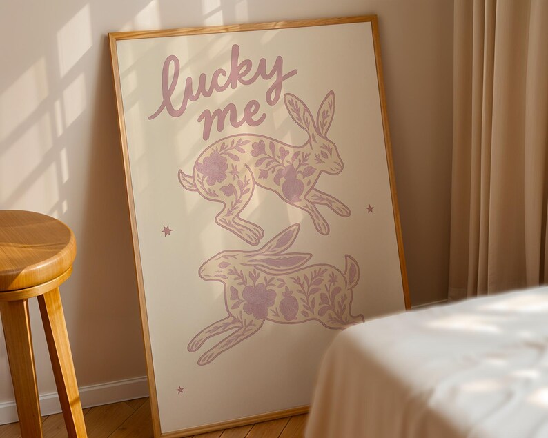 May include: A framed art print featuring two running rabbits with floral patterns in a soft purple hue. The words "lucky me" are written above the rabbits in a cursive font. The background is a light beige color.