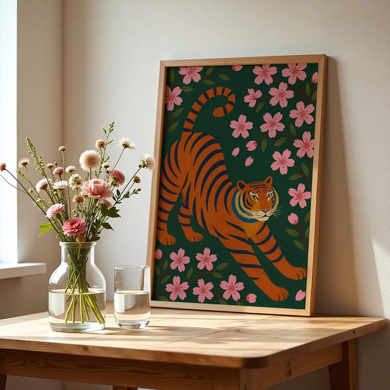 May include: A framed art print featuring an orange tiger with blue stripes, set against a dark green background with pink cherry blossoms. The artwork is displayed on a wooden table, next to a vase of flowers and a glass of water.