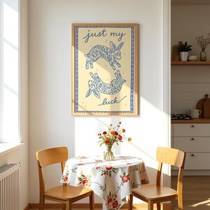 May include: A framed art print featuring two blue rabbit illustrations with floral patterns on a cream background. The text "just my luck" is written in cursive. The print is displayed above a small table with a floral tablecloth.
