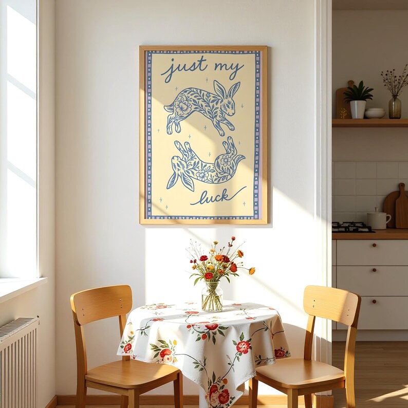 May include: A framed art print featuring two blue rabbit illustrations with floral patterns on a cream background. The text "just my luck" is written in cursive. The print is displayed above a small table with a floral tablecloth.