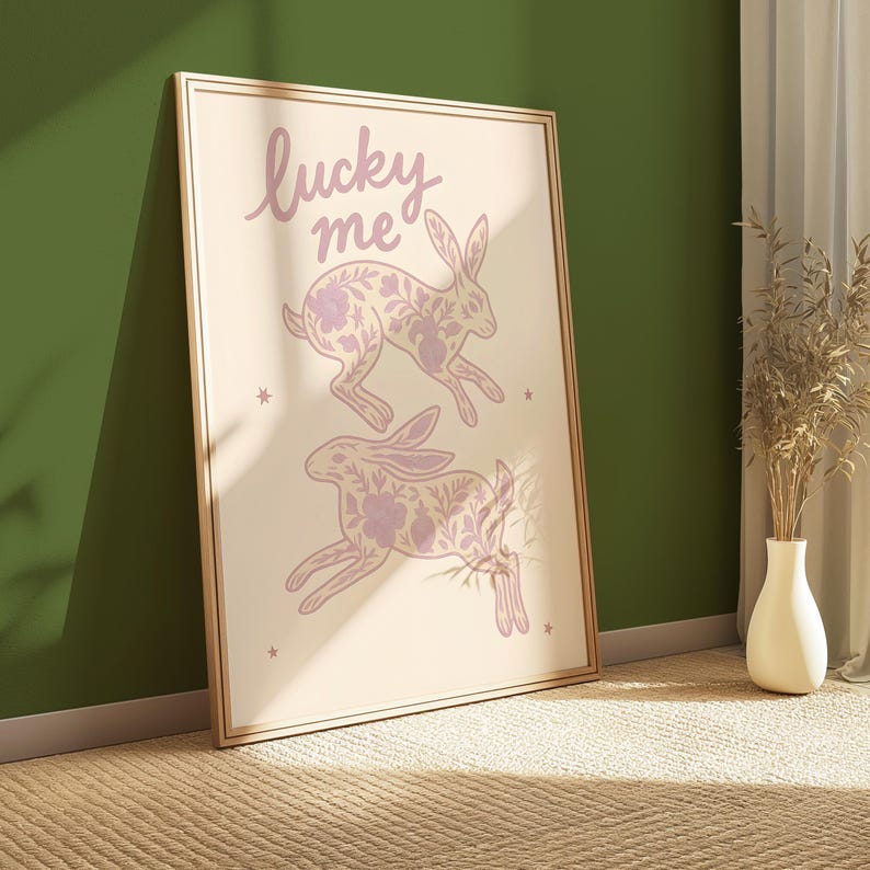 May include: A framed art print featuring two leaping rabbits with floral patterns in light purple. The words "lucky me" are written above the rabbits in a cursive font. The print is set against a cream background with small star accents.