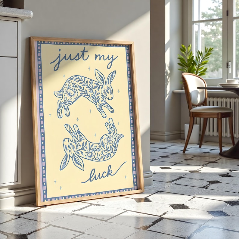 May include: A framed art print featuring two blue rabbit illustrations with floral patterns on a light yellow background. The words "just my luck" are written in blue script. The print has a light wood frame and a blue border.