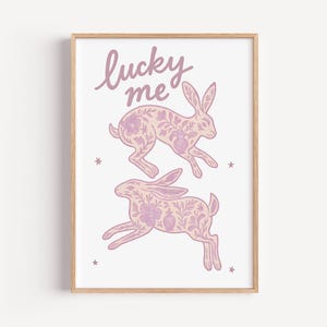 May include: A framed art print featuring two leaping rabbits with floral patterns in shades of pink and lavender. The words "lucky me" are written above the rabbits in a cursive font. Small star shapes are scattered around the rabbits.