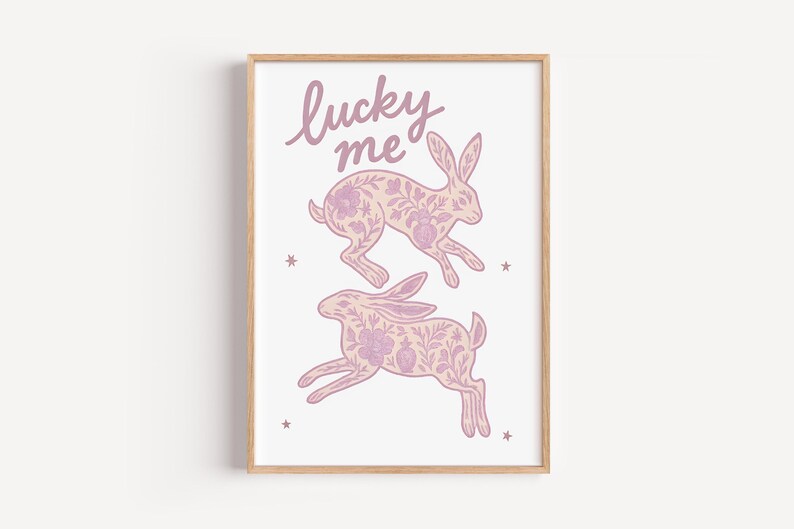 May include: A framed art print featuring two leaping rabbits with floral patterns in shades of pink and lavender. The words "lucky me" are written above the rabbits in a cursive font. Small star shapes are scattered around the rabbits.
