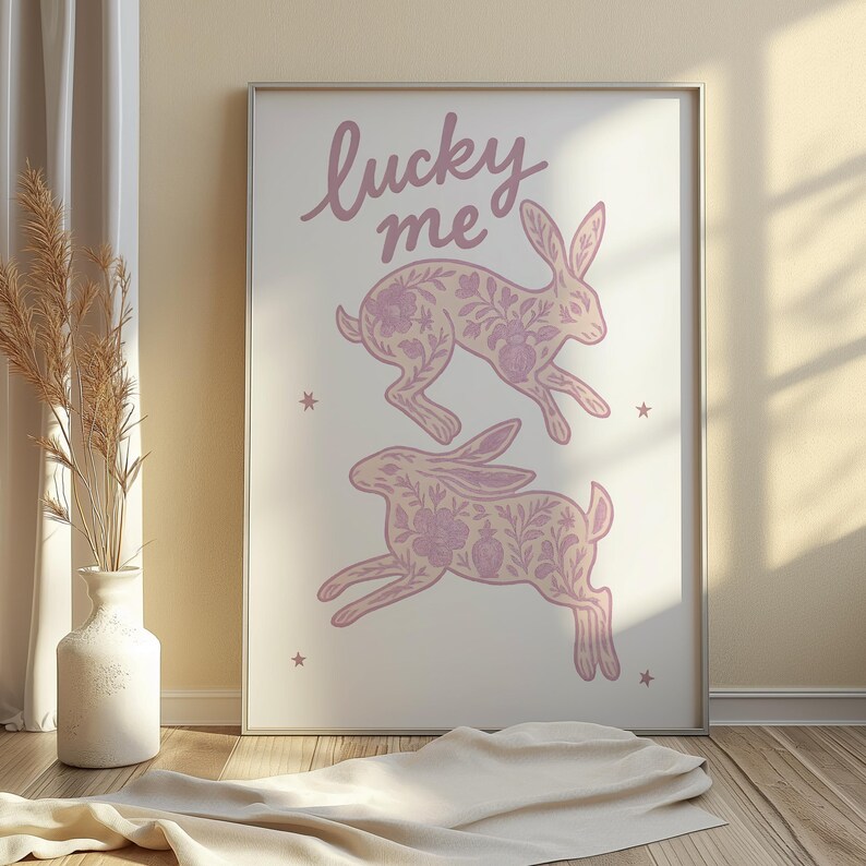May include: A framed print featuring two leaping rabbits with floral patterns in a soft purple hue. The words "lucky me" are written above the rabbits in a cursive font. The print is on a cream-colored background.