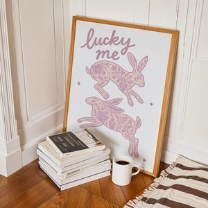 May include: A framed art print featuring two leaping rabbits with floral patterns in shades of lavender. The words "lucky me" are written above the rabbits. A stack of books and a white mug with coffee are in the foreground.