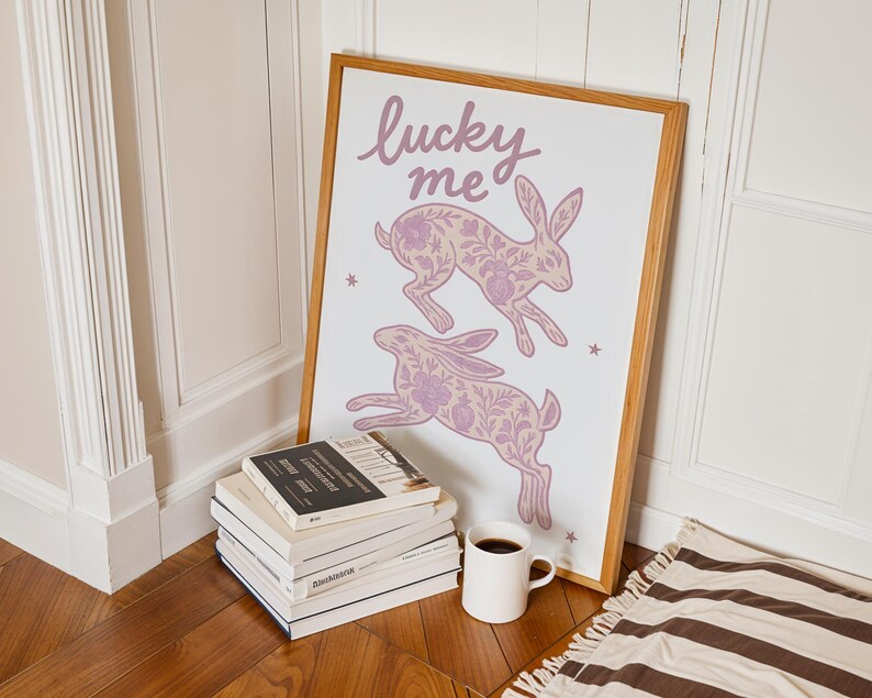 May include: A framed art print featuring two leaping rabbits with floral patterns in shades of lavender. The words "lucky me" are written above the rabbits. A stack of books and a white mug with coffee are in the foreground.