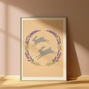 May include: Framed art print with two grey hares running within a wreath of lavender and green leaves. The artwork has a neutral colour palette, with a beige background and a white frame.