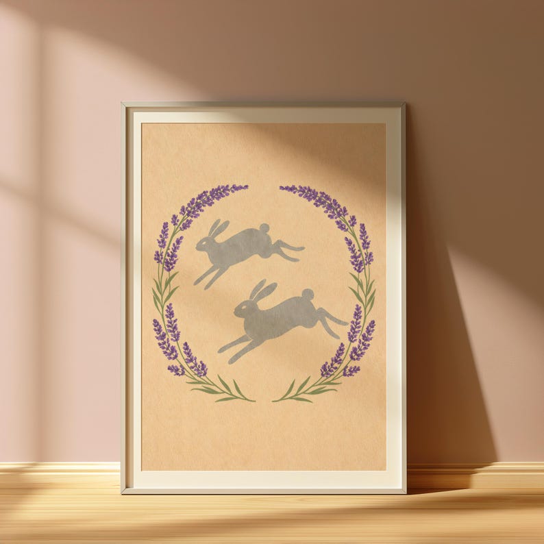 May include: Framed art print with two grey hares running within a wreath of lavender and green leaves. The artwork has a neutral colour palette, with a beige background and a white frame.