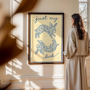 May include: A framed art print featuring two blue floral rabbit illustrations on a light yellow background. The words "just my luck" are written in cursive. The frame is dark brown.
