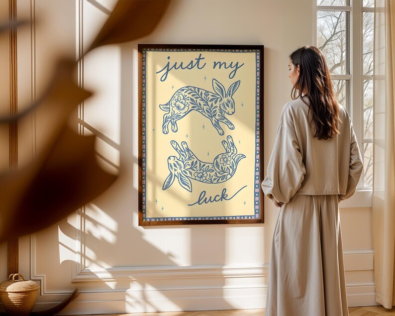 May include: A framed art print featuring two blue floral rabbit illustrations on a light yellow background. The words "just my luck" are written in cursive. The frame is dark brown.