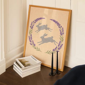 May include: A framed print with two grey rabbits within a lavender and green wreath. The artwork is on a wooden floor, beside a stack of magazines and two black candlesticks.
