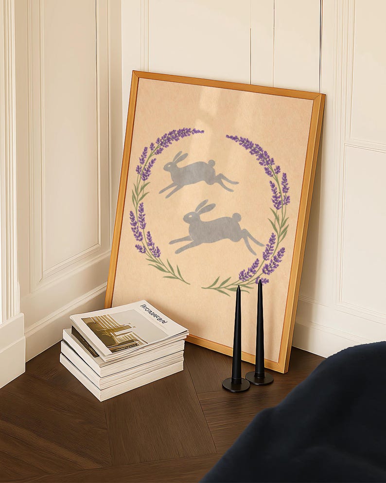 May include: A framed print with two grey rabbits within a lavender and green wreath. The artwork is on a wooden floor, beside a stack of magazines and two black candlesticks.