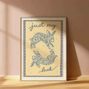 May include: Framed art print featuring two stylized rabbits with floral patterns, one upright and one inverted. The print has a light yellow background with the words "just my luck" in cursive. The frame is a light color with a decorative border.