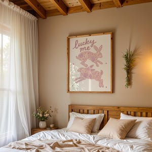 May include: A bedroom scene featuring a wooden bed frame with white and peach-colored bedding. A framed print with two rabbits and the text "lucky me" hangs above the bed. Sheer white curtains frame a window, and a hanging light fixture is visible.