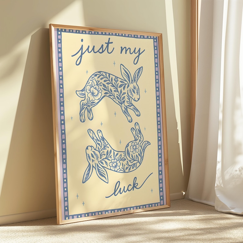 May include: A framed art print featuring two blue floral rabbit illustrations on a cream background. The words "just my luck" are written in blue script. The print has a light brown wooden frame and a blue and purple border.