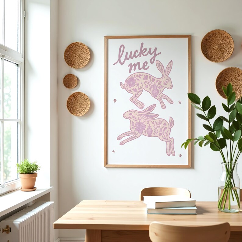 May include: A framed print with the words "lucky me" and two pink rabbits with floral patterns. The print is displayed on a white wall, alongside woven wall decor and a dining table. A vase of greenery sits on the table.
