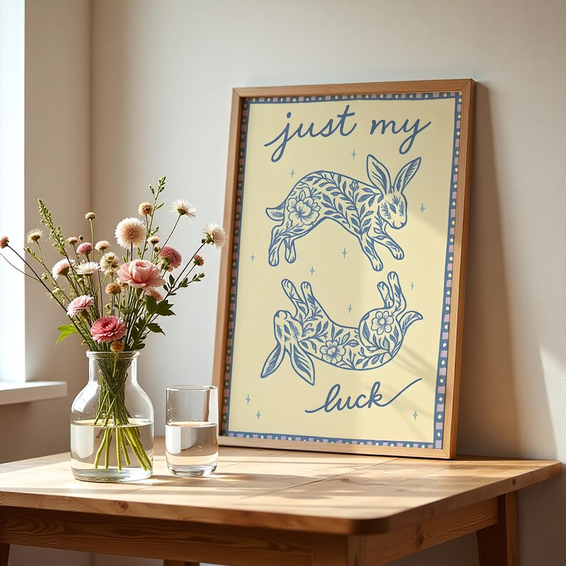 May include: A framed art print featuring two blue floral rabbit illustrations on a light yellow background. The text "just my luck" is written in cursive. The print is displayed on a wooden table with a vase of flowers.
