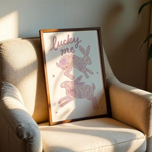 May include: A framed art print featuring two pink rabbits with floral patterns, and the words "lucky me" in cursive. The artwork is displayed on a beige armchair with a wooden frame.