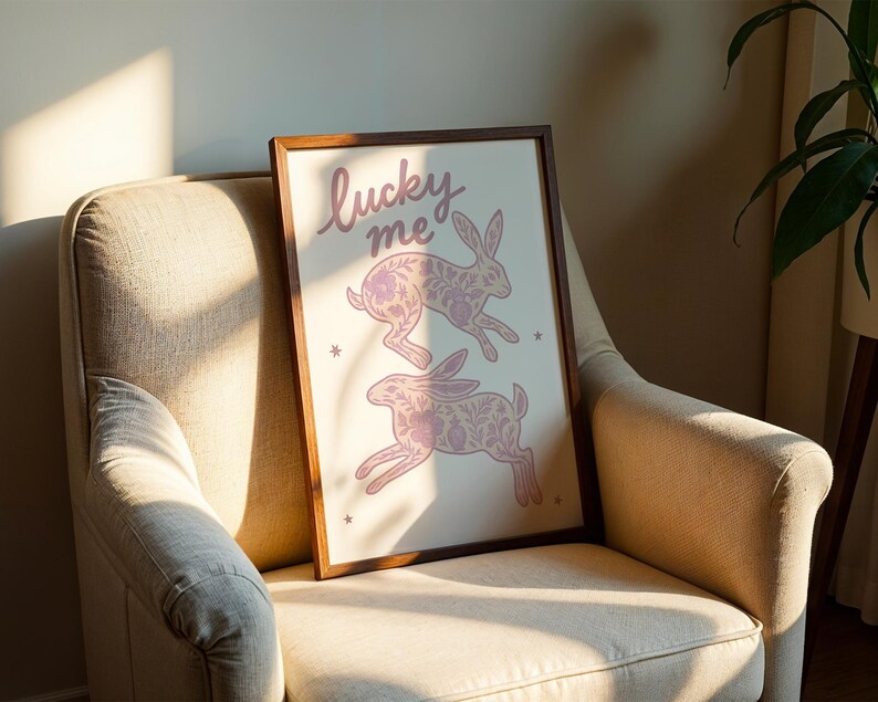 May include: A framed art print featuring two pink rabbits with floral patterns, and the words "lucky me" in cursive. The artwork is displayed on a beige armchair with a wooden frame.