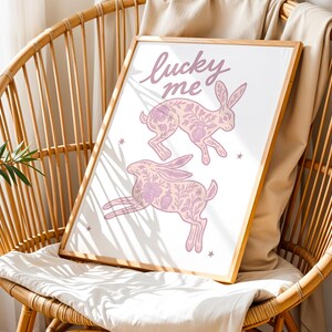 May include: A framed print featuring two pink rabbits with floral patterns and the text "lucky me" in cursive. The artwork is set against a white background and framed in a light wood. The print is displayed on a wicker chair with a white cushion.