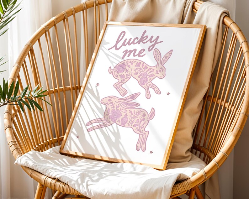 May include: A framed print featuring two pink rabbits with floral patterns and the text "lucky me" in cursive. The artwork is set against a white background and framed in a light wood. The print is displayed on a wicker chair with a white cushion.