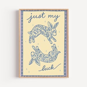 May include: A framed art print featuring two blue rabbits with floral patterns on a light yellow background. The words "just my luck" are written in cursive. The print has a light brown frame and a decorative border.