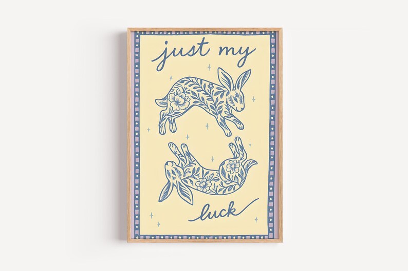 May include: A framed art print featuring two blue rabbits with floral patterns on a light yellow background. The words "just my luck" are written in cursive. The print has a light brown frame and a decorative border.