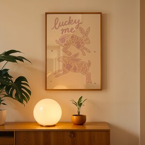 May include: A framed print with two pink rabbits and the text "lucky me" hangs on a wall. A glowing orb lamp sits on a wooden cabinet, with a potted plant to the left and a small plant in a terracotta pot to the right.