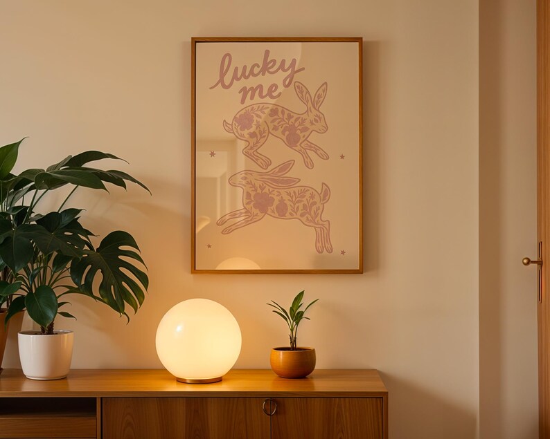 May include: A framed print with two pink rabbits and the text "lucky me" hangs on a wall. A glowing orb lamp sits on a wooden cabinet, with a potted plant to the left and a small plant in a terracotta pot to the right.