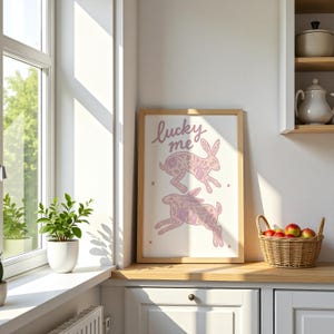 May include: A framed print featuring two pink rabbits with floral patterns, and the text "lucky me". The artwork is displayed on a wooden countertop in a kitchen setting, with a window and a basket of apples nearby.