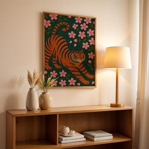 May include: A framed art print featuring an orange tiger with black stripes, set against a dark green background with pink cherry blossoms. The artwork is displayed above a wooden shelf with decorative vases and a lamp.