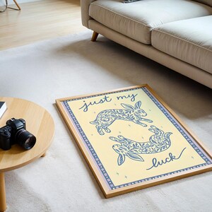 May include: A framed art print featuring two stylized rabbits with floral patterns in blue on a yellow background. The text "just my luck" is written in cursive. The frame is light brown.