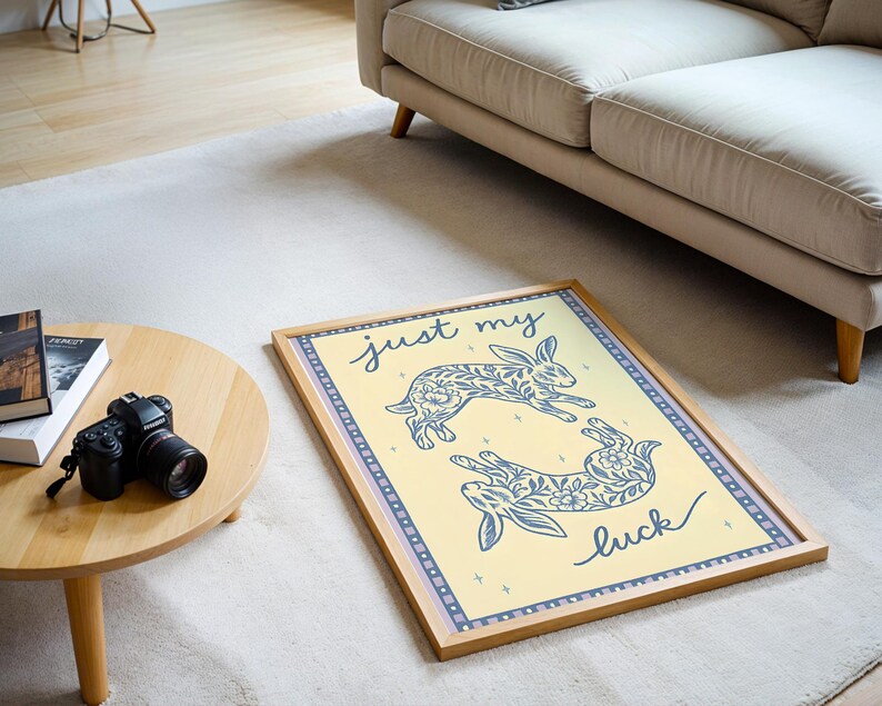 May include: A framed art print featuring two stylized rabbits with floral patterns in blue on a yellow background. The text "just my luck" is written in cursive. The frame is light brown.