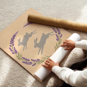 May include: A rolled print featuring two grey rabbit silhouettes within a lavender and green floral wreath. The print is on a tan background. A rolled white print and a brown tube are also visible.
