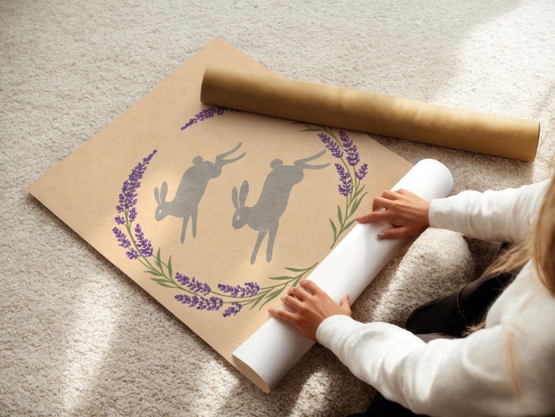 May include: A rolled print featuring two grey rabbit silhouettes within a lavender and green floral wreath. The print is on a tan background. A rolled white print and a brown tube are also visible.
