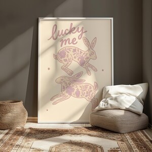 May include: A framed art print featuring two running rabbits with floral patterns in a soft purple hue. The words "lucky me" are written above the rabbits in a matching color. The background is a light beige.